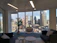 A modern office space with large floor-to-ceiling windows offering a panoramic view of a city skyline, including several skyscrapers. The interior features contemporary furniture including dark sofas, a round glass hanging chair, and a small table. A potted plant adds a natural element near the window.