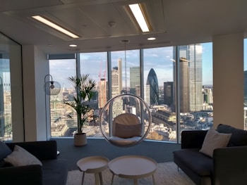 A modern office space with large floor-to-ceiling windows offering a panoramic view of a city skyline, including several skyscrapers. The interior features contemporary furniture including dark sofas, a round glass hanging chair, and a small table. A potted plant adds a natural element near the window.