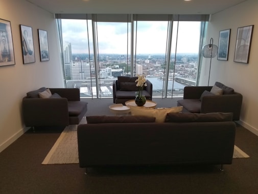 A modern, stylish living room with large floor-to-ceiling windows offering a panoramic city view. The room is furnished with dark gray sofas arranged around a small coffee table with a potted plant at the center. The walls are adorned with minimalist artwork and a standing floor lamp provides additional lighting.