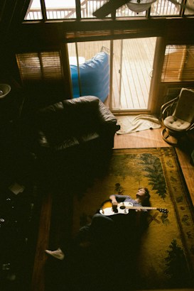 An overhead view of a cozy room with wooden accents and a patterned rug. A person is lying on the rug, holding an acoustic guitar. Soft light filters through a glass door leading to a wooden deck, creating a mellow atmosphere.