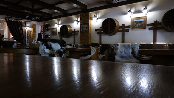 The interior of a cozy restaurant or bar is depicted, featuring wooden furniture and rustic decor. The walls are adorned with round wooden barrel lids and a couple of framed pictures. Several chairs and benches are covered with animal furs, enhancing the rustic ambiance. The lighting is warm and soft, provided by wall-mounted lamps that create a welcoming atmosphere.