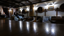 The interior of a cozy restaurant or bar is depicted, featuring wooden furniture and rustic decor. The walls are adorned with round wooden barrel lids and a couple of framed pictures. Several chairs and benches are covered with animal furs, enhancing the rustic ambiance. The lighting is warm and soft, provided by wall-mounted lamps that create a welcoming atmosphere.