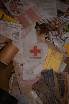 A collection of scattered papers and documents, including a prominent leaflet from the Red Cross with the logo and text, old newspapers, booklets, and other printed materials. Some items appear worn and aged, hinting at neglect or disorder.