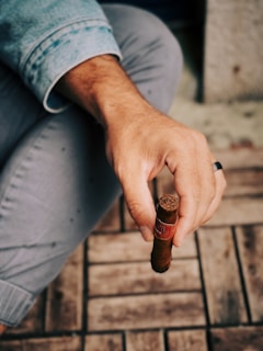 A person is holding a cigar in their hand, with a focus on the fingers and the cigar. The individual is wearing a light blue denim jacket, visible on the wrist, and a silver ring on the index finger. The background consists of a wooden tiled surface.