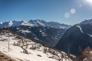 A picturesque mountain landscape featuring snow-covered peaks under a clear blue sky. Small houses and winding roads are scattered across the snow-laden hillside.