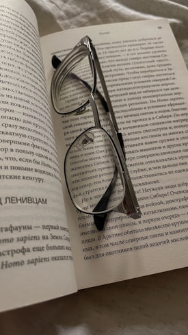 A pair of reading glasses rests open on the pages of a book filled with text in a non-Latin script, likely Cyrillic. The book is lying on a soft surface, possibly a bed or couch. The glasses are silver with black earpieces, and they are placed across the pages, partially obscuring the text.