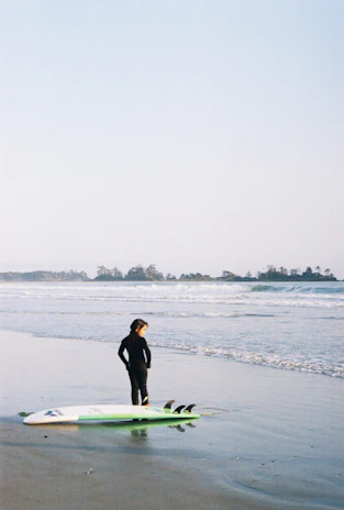 A person in a wetsuit stands on a sandy beach next to a surfboard, gazing towards the ocean. Waves gently crash onto the shore, and tree-lined islands are visible in the distance. The scene is calm and serene with a soft, natural light.