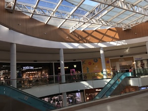 A spacious shopping mall interior with a glass skylight allowing natural light to illuminate the area. Several shops are visible, including a clothing store. There is an escalator on the right side. The architecture features curved walls and multiple support columns.