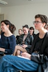 A group of people is seated in a bright room, focusing on a presentation or speaker. They appear engaged and slightly amused. The setting suggests a classroom or workshop environment. The individuals present are casually dressed and are taking notes.