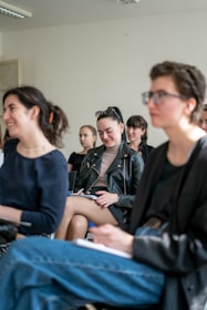 A group of people is seated in a bright room, focusing on a presentation or speaker. They appear engaged and slightly amused. The setting suggests a classroom or workshop environment. The individuals present are casually dressed and are taking notes.