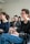 A group of people is seated in a bright room, focusing on a presentation or speaker. They appear engaged and slightly amused. The setting suggests a classroom or workshop environment. The individuals present are casually dressed and are taking notes.