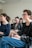 A group of people is seated in a bright room, focusing on a presentation or speaker. They appear engaged and slightly amused. The setting suggests a classroom or workshop environment. The individuals present are casually dressed and are taking notes.
