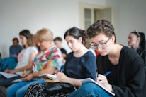 A group of people sitting in chairs, focusing on taking notes. The individuals appear engaged, with some looking down at their notebooks or writing carefully.