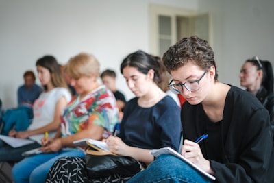 A group of people sitting in chairs, focusing on taking notes. The individuals appear engaged, with some looking down at their notebooks or writing carefully.