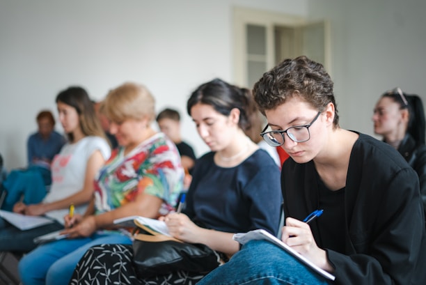 A group of people sitting in chairs, focusing on taking notes. The individuals appear engaged, with some looking down at their notebooks or writing carefully.