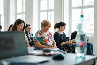 A group of people seated in a bright room with large windows, each focused on taking notes. In the foreground, there is a blurred laptop and a water bottle, suggesting an educational or workshop setting.