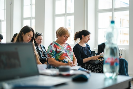 A group of people seated in a bright room with large windows, each focused on taking notes. In the foreground, there is a blurred laptop and a water bottle, suggesting an educational or workshop setting.