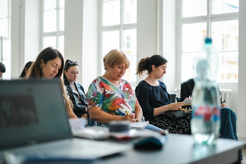 A group of people seated in a bright room with large windows, each focused on taking notes. In the foreground, there is a blurred laptop and a water bottle, suggesting an educational or workshop setting.
