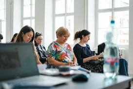A group of people seated in a bright room with large windows, each focused on taking notes. In the foreground, there is a blurred laptop and a water bottle, suggesting an educational or workshop setting.