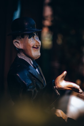 A figurine of a man wearing a black suit, white shirt, and a bowler hat. The figure has exaggerated facial features and is positioned with one hand extended as if gesturing. The background is blurred and dark, giving prominence to the figurine.