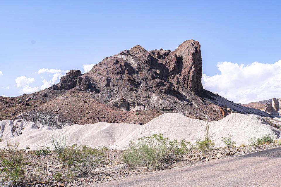 The image depicts a rocky mountain landscape with a large, jagged rock formation on the right side and a smaller, smoother rock formation on the left side. The mountains are covered in dirt and rocks, creating a rugged terrain. In the foreground, there is a road running through the scene, adding an element of human presence to the natural setting.

The sky above is clear blue with scattered clouds, providing a beautiful backdrop for the rocky landscape below.