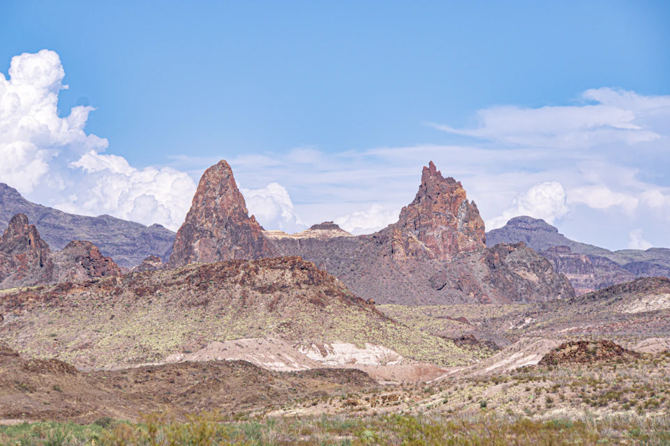 The image depicts a breathtaking view of two towering red rock formations, known as "the Big Rock" and "Big Pine," situated on a vast desert plain. The rock formations are covered in patches of green vegetation, adding a touch of color to the otherwise monochrome landscape. The sky above is a clear blue with scattered white clouds, creating a striking contrast against the red rocks below.