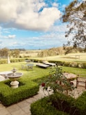 A serene garden setting with well-manicured hedges and a central stone fountain. Lawn chairs and a metal garden table are set on the lush green grass. Wooden patio furniture, including a bench and a round table, complement the outdoor area. Red flowers bloom in a bush nearby, and the garden opens up to a vast, scenic landscape with trees under a sky filled with fluffy white clouds.