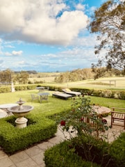 A serene garden setting with well-manicured hedges and a central stone fountain. Lawn chairs and a metal garden table are set on the lush green grass. Wooden patio furniture, including a bench and a round table, complement the outdoor area. Red flowers bloom in a bush nearby, and the garden opens up to a vast, scenic landscape with trees under a sky filled with fluffy white clouds.