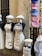 Mannequins are dressed in white aprons and chef hats with embroidered names and phrases. To the right, a display of colorful caps with embroidered names is attached to a wall. The setting appears to be an outdoor street shop, with stone walls and racks of merchandise.