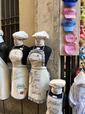 Mannequins are dressed in white aprons and chef hats with embroidered names and phrases. To the right, a display of colorful caps with embroidered names is attached to a wall. The setting appears to be an outdoor street shop, with stone walls and racks of merchandise.