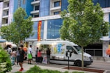 A food truck is parked on the side of a street, with several people gathered around it, placing orders. The truck features brightly colored signage showcasing various food items. Two trees are prominently situated on the pavement nearby, with a modern building in the background partially under construction.