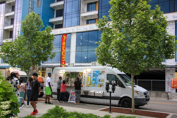 A food truck is parked on the side of a street, with several people gathered around it, placing orders. The truck features brightly colored signage showcasing various food items. Two trees are prominently situated on the pavement nearby, with a modern building in the background partially under construction.