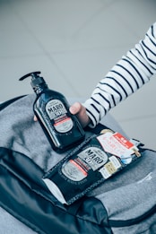 A close-up view of two hair product containers set on top of a gray bag. One is a pump bottle being held by someone wearing a striped shirt, while the other is a refill pouch with printed labels and Japanese text.