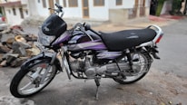 A black and purple motorcycle is parked on a roadside. The motorcycle displays visible signs of wear, with scratches and a torn seat. There is a pile of rocks and a residential building with a white exterior in the background.