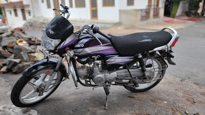 A black and purple motorcycle is parked on a roadside. The motorcycle displays visible signs of wear, with scratches and a torn seat. There is a pile of rocks and a residential building with a white exterior in the background.