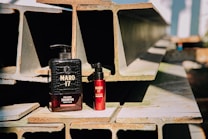 Two bottles of Maro 17 collagen shampoo and hair product are placed on a stack of industrial metal beams. The large black bottle contrasts with a smaller red bottle next to it. The scene is well-lit with natural sunlight casting shadows, highlighting the texture of the beams.