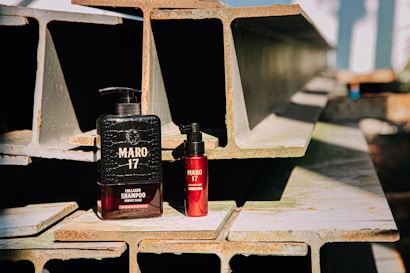 Two bottles of Maro 17 collagen shampoo and hair product are placed on a stack of industrial metal beams. The large black bottle contrasts with a smaller red bottle next to it. The scene is well-lit with natural sunlight casting shadows, highlighting the texture of the beams.