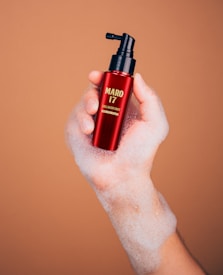 A hand covered in soap suds holds a red bottle with a black pump dispenser, labeled 'MARO 17 COLLAGEN SHOT,' against a light brown background.