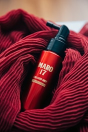 A red bottle labeled 'MARO 17 COLLAGEN SHOT' is nestled in a soft, textured red fabric. The bottle features a pump top, and the vibrant red of the fabric complements the glossy finish of the bottle.