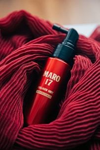 A red bottle labeled 'MARO 17 COLLAGEN SHOT' is nestled in a soft, textured red fabric. The bottle features a pump top, and the vibrant red of the fabric complements the glossy finish of the bottle.