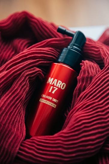 A red bottle labeled 'MARO 17 COLLAGEN SHOT' is nestled in a soft, textured red fabric. The bottle features a pump top, and the vibrant red of the fabric complements the glossy finish of the bottle.