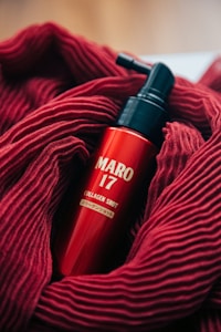 A red bottle labeled 'MARO 17 COLLAGEN SHOT' is nestled in a soft, textured red fabric. The bottle features a pump top, and the vibrant red of the fabric complements the glossy finish of the bottle.