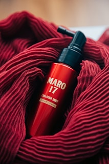 A red bottle labeled 'MARO 17 COLLAGEN SHOT' is nestled in a soft, textured red fabric. The bottle features a pump top, and the vibrant red of the fabric complements the glossy finish of the bottle.