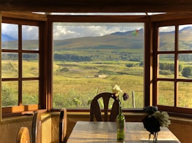 A scenic view is visible through a wide wooden window, showcasing a vast expanse of rolling green hills under a cloudy sky. Inside the room, a wooden table with two chairs is adorned with a wine bottle holding white and dark flowers.