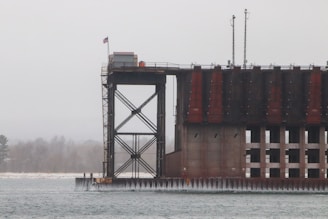 A large industrial structure, likely a dam or lock, is situated next to a body of water. The structure displays a series of vertical steel beams and concrete elements, with a small American flag visible at the top. The scene is overcast, with a misty or foggy backdrop that obscures distant trees.