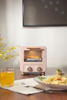 A small, pastel-colored toaster oven is placed on a wooden table, with toast inside it. Bright yellow flowers are arranged in the background, next to a framed artwork on the wall. In the foreground, there is a glass of orange juice and a plate of spaghetti garnished with greens and a tomato slice.