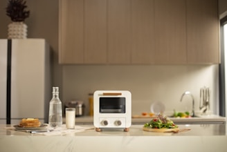 A modern kitchen countertop features a small white oven at the center. On the left side, there is a stack of toast with a bottle of water and a glass of milk nearby. On the right, a fresh green salad is placed on a wooden plate. Background elements include a sleek refrigerator, wooden cabinets, and a sink with a tap.