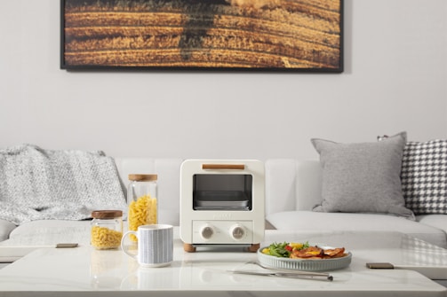 A minimalistic living room setup features a white couch adorned with textured gray and patterned cushions. In front of the couch, a modern coffee table holds a small white toaster oven. Next to the oven, there are two glass jars filled with pasta, and a mug with a geometric pattern. A plate of food, a fork, and a smartphone also rest on the table. A large photograph of a field hangs on the wall behind the couch.