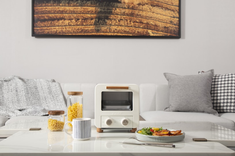 A minimalistic living room setup features a white couch adorned with textured gray and patterned cushions. In front of the couch, a modern coffee table holds a small white toaster oven. Next to the oven, there are two glass jars filled with pasta, and a mug with a geometric pattern. A plate of food, a fork, and a smartphone also rest on the table. A large photograph of a field hangs on the wall behind the couch.