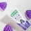 A tube of G.U.M toothpaste with grape flavor is positioned on a light surface. The packaging features purple and green accents, displaying information about cavity prevention and suitability for young children. Surrounding the toothpaste are plastic grape-shaped objects in a vibrant purple color.